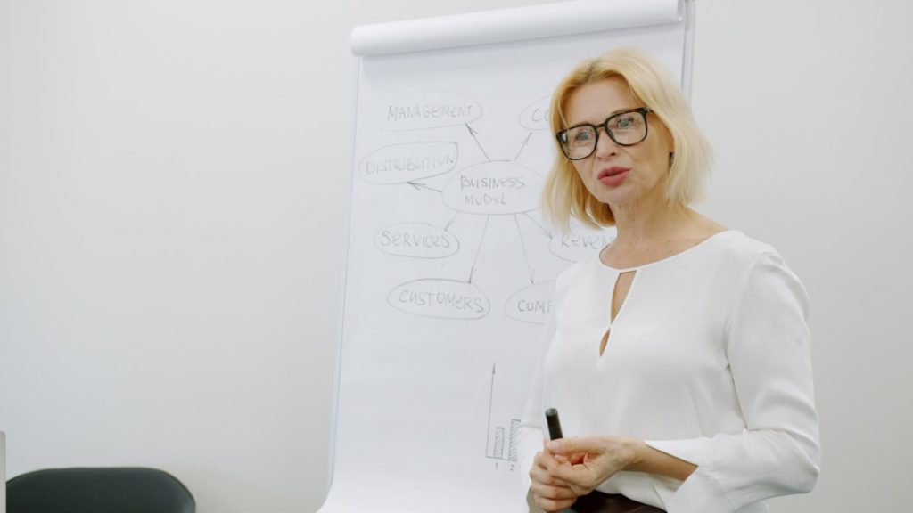 Mature businesswoman in elegant clothing is speaking near flipchart board explaining business and marketing in office. People and analytics concept.